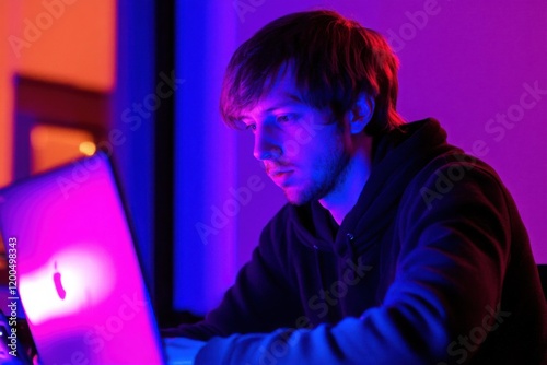 Young man focused on his laptop with vibrant purple and blue lighting, showcasing modern technology use, creativity, and a relaxed nighttime environment at home. photo