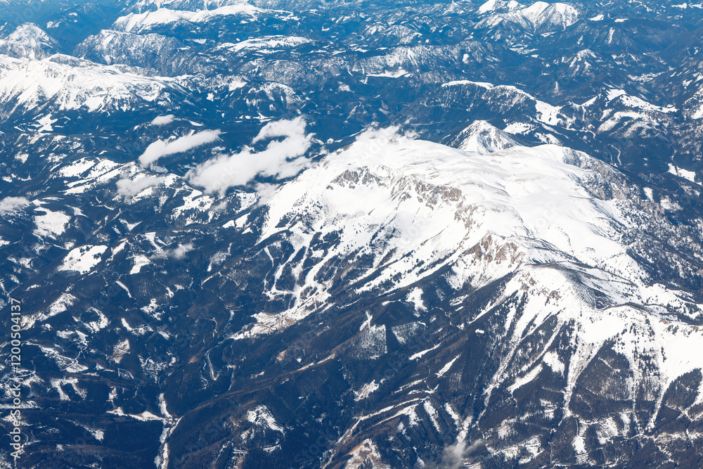 Aerial view of a mountainous region with snow-covered peaks and ridges. The landscape features a mix of snow and dark forested areas, with some clouds partially covering the mountains