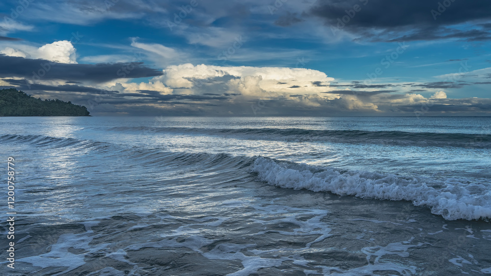 Obraz premium Morning. The clouds in the blue sky are highlighted in gold. The ocean waves roll towards the shore, foam, spread out. Glare on the sea surface. A hill in the distance. Malaysia. Borneo. Kota Kinabalu