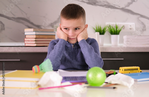 Frustrated, unhappy child sits down at table and does his homework among stack of books and scattered pieces of paper and toys. Education school, learning disabilities, reading difficulties, dyslexia.