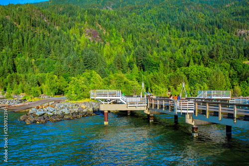 Walking pier in Porteau Cove, a view from the sea, three  people, blue water against the backdrop of a mountain slope overgrown with coniferous forest and deciduous trees on the shore photo