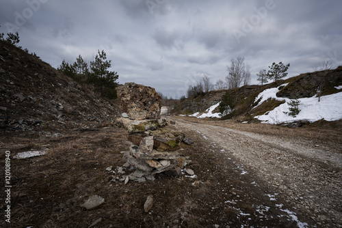 Abandoned quarry in spring: dirt road with tire tracks, remnants of snow, stone pyramid and cloudy sky.