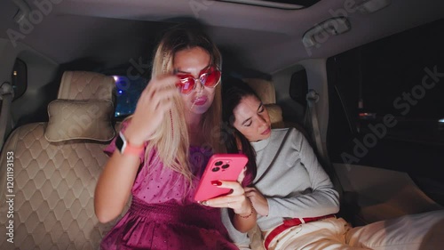 Two friends ride home in a taxi at night after a party. One is on her phone, while the other drunkenly sleeps on her shoulder. photo