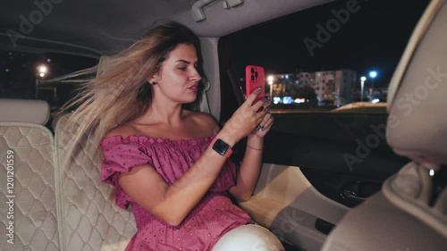 A blonde girl rides home in a taxi at night, capturing the beautiful city lights with her smartphone. She enjoys the mesmerizing view of the nighttime city.