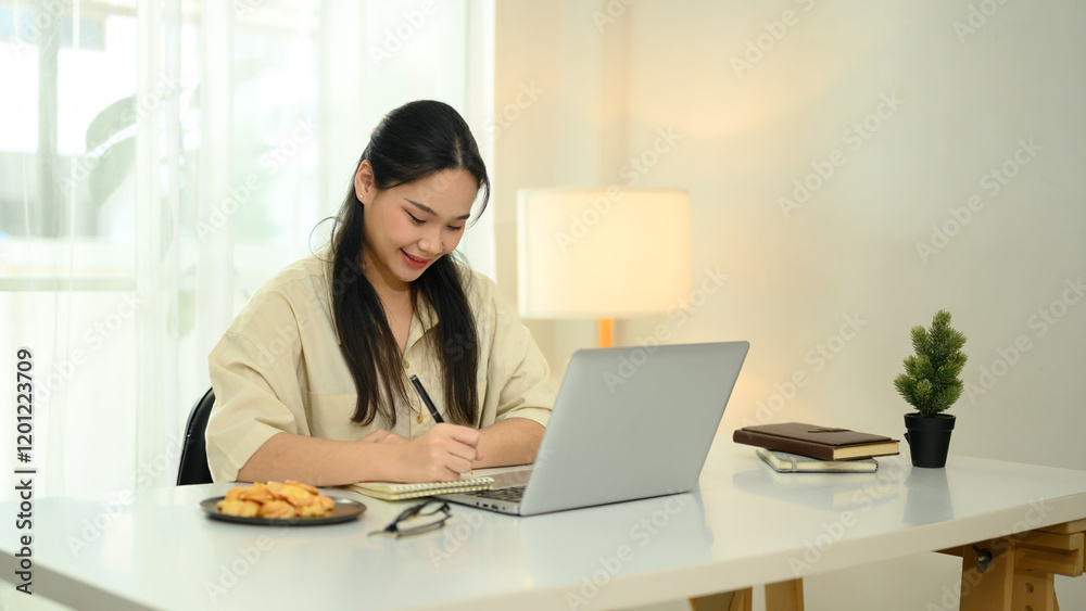 Smiling young woman writing in a notebook while working on her laptop Smiling young woman writing in a notebook while working on her laptop