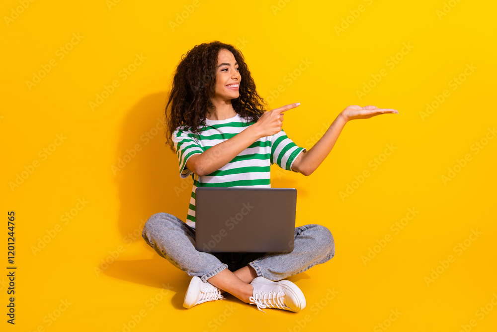 Full body portrait of nice young girl laptop direct finger hold empty space wear striped t-shirt isolated on yellow color background Full body portrait of nice young girl laptop direct finger hold empty space wear striped t-shirt isolated on yellow color background