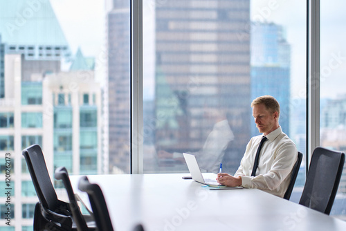Busy businessman financial analyst or manager using computer technology at work. Young business man executive wearing tie working on laptop writing notes in skyscraper office at desk near big window.