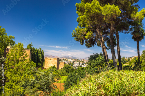 Alhambra complex in Granada town, Andalusia, Spain. Serene view of a city nestled in mountains, seen from a lush garden with ancient structures.