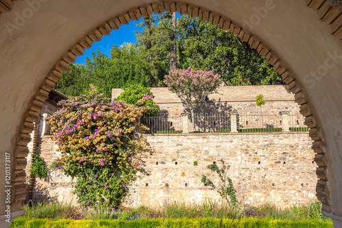 Alhambra complex in Granada town, Andalusia, Spain. Stone archway view of a garden with flowering plants and a stone wall.