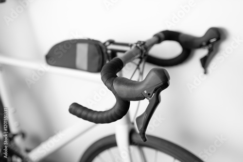 A bicycle with a black handlebar and a black bag on the handlebar. The bag is a black and white backpack. The bike is parked against a white wall photo