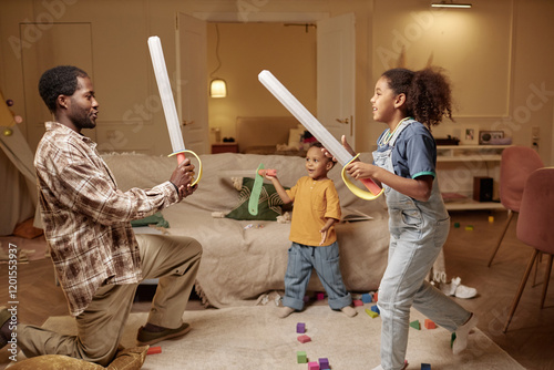 Side view of African American father playing with children fighting with toy swords, while reciting lines of noble knight standing on one knee in messy living room photo