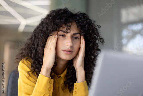 Headache, woman is overloaded and overtired at workplace inside office working with laptop. Office worker holds head in hands, looking at computer screen. photo