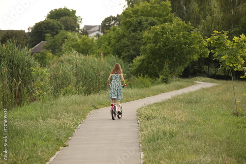A girl rides a bicycle on an asphalt avenue among the greenery of the park. Active fun outdoors. Sports load for the proper development of the girl's body.