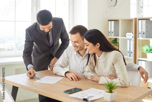 Confident young male realtor agent pointing at paper for family couple of customers, clients signing, giving consultation, legal help with mortgage, real estate property buying, contract agreement