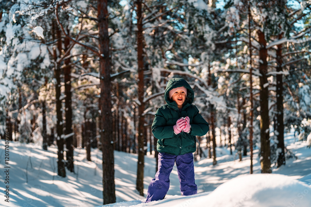 Naklejka premium Smiling child dressed in warm winter clothing is joyfully crafting a snowball in a snowy forest, embracing the fun of winter holidays amidst the stunning natural landscape