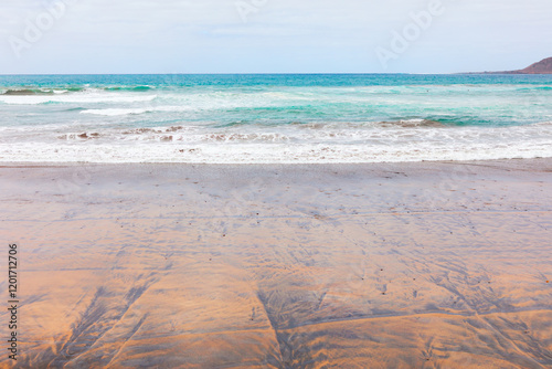 Obraz na płótnie Serene view of the Pacific coast featuring a sandy beach with intricate patterns, gentle waves rolling onto the shore, and turquoise ocean waters meeting the horizon under a soft, cloudy sky w salonie
