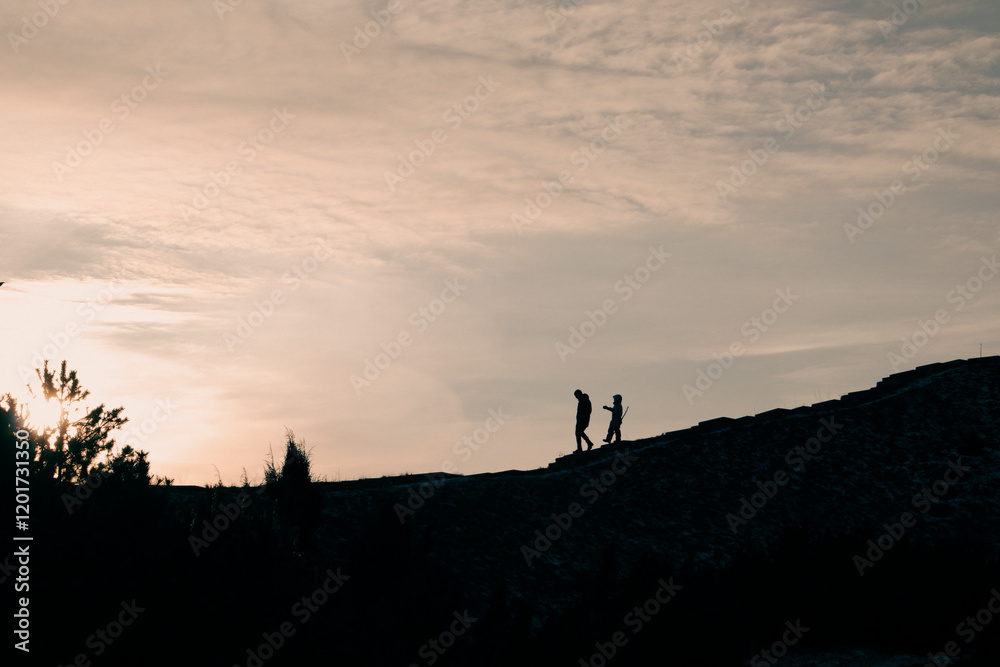 Fototapeta premium Contrasting silhouettes of two people in sunset light - an adult and a kid - on a hill in a park, forest. Family nature walk with a child. Adventure.