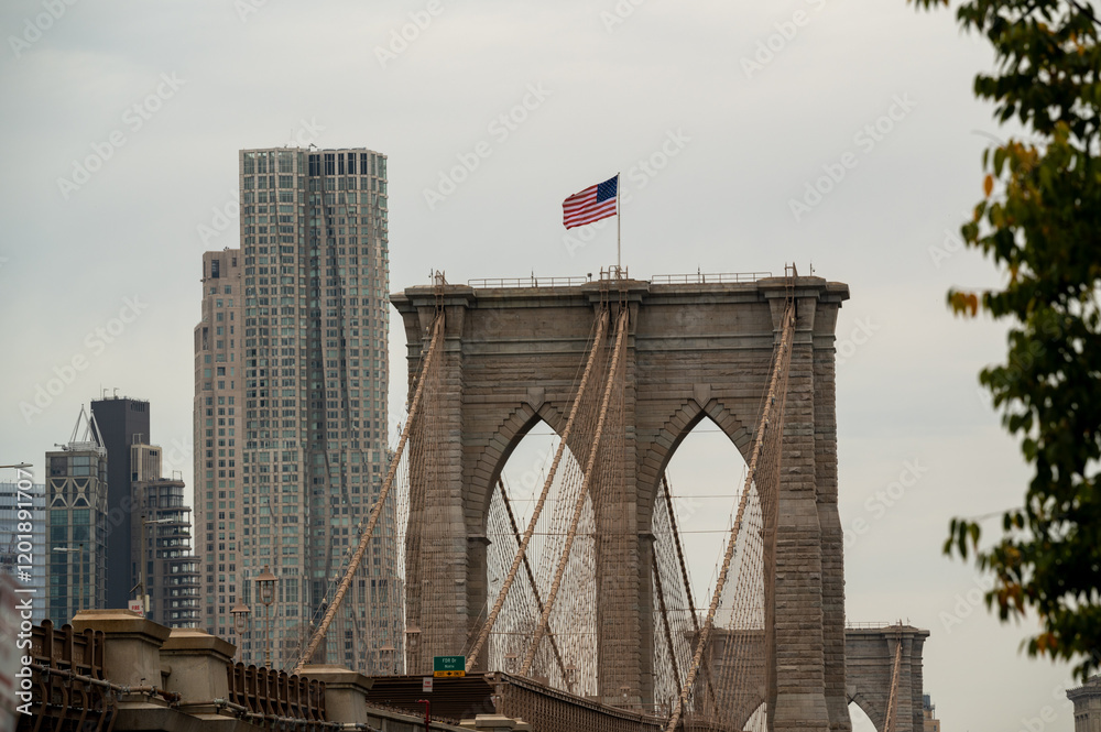 Fototapeta premium Brooklyn Bridge viewed from below on an overcast day with the American flag waving against a backdrop of city buildings