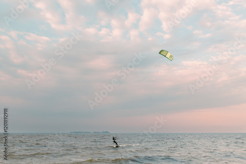 Kitesurfer riding on waves with green kite at sunset over open sea. Dynamic water sports scene with pastel sky in background