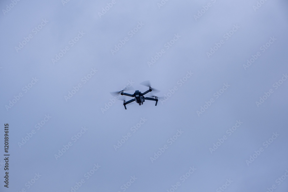 Black quadcopter drone flies in gray winter sky. Propellers are blurred in motion as it hovers steadily in air.