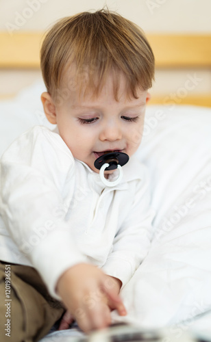 Child playing quietly on a bed. A young child with a pacifier explores a book while resting on a soft bed in a cozy room.