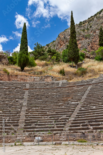 Generated imagThe ancient theater of Delphi, with stone seating arranged in semicircles, surrounded by cypress trees and rocky hills under a bright blue sky.e
