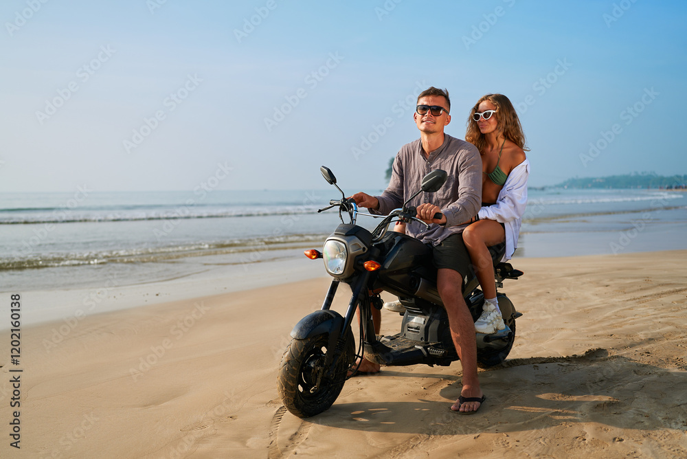 Obraz premium Couple rides motorbike on sandy beach near ocean. Man drives motorcycle, woman sits behind smiling. Enjoying adventure travel, seaside freedom, summer vibes.