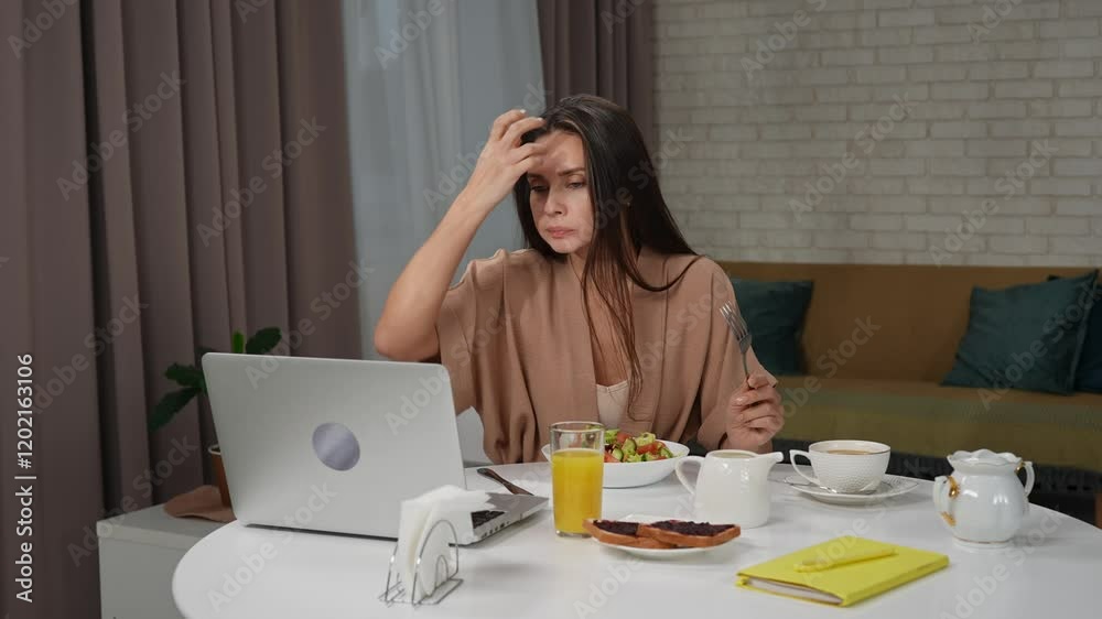 Young brunette woman in the morning in the living room at home enjoying healthy breakfast, sitting at table eating salad and checking work on laptop. Young brunette woman in the morning in the living room at home enjoying healthy breakfast, sitting at table eating salad and checking work on laptop.