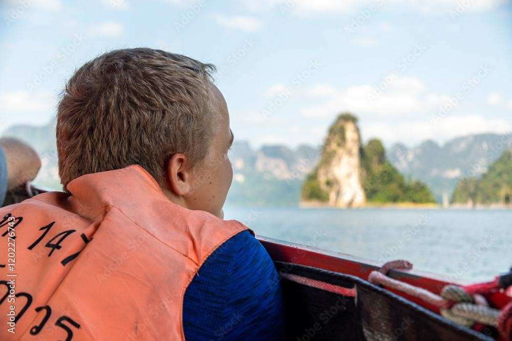 Fototapeta premium Smiling young boy wearing life jacket on boat in calm waters surrounded by mountains during cloudy day