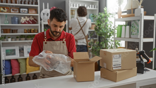 Men working together in a home decor shop, one packing items carefully in boxes while the other arranges shelves in the store interior. photo