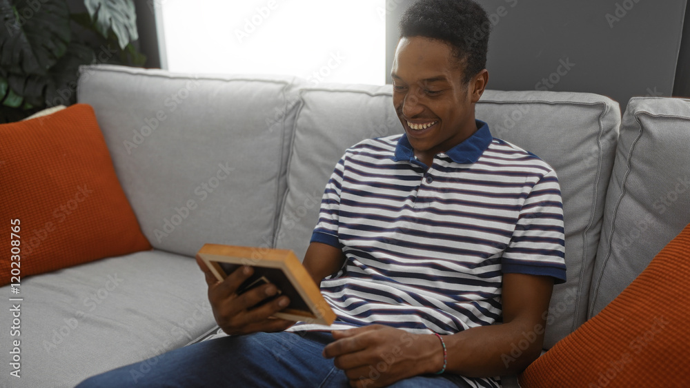 Fototapeta premium Young man smiling while sitting on a couch in a cozy living room holding a photo frame, showcasing a comfortable indoor environment filled with modern interior design elements.