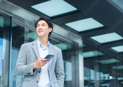 Portrait of attractive young asian businessman standing in front of a modern office building. Smiling Asian executive using smartphone for business, working, texting a message in city street outdoors.