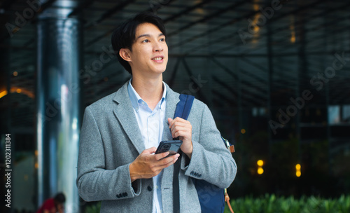 Successful young Asian businessman using a smartphone and, standing in front of a modern office building. Confident Smiling asian man worker looking around while walking on city street outdoors.