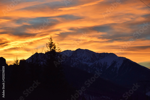 Golden hues of the sunset sky, blending vibrant shades of orange and yellow, cast a warm glow over the majestic High Tatras mountain peaks. Dark silhouettes of trees, dramatic contrast.