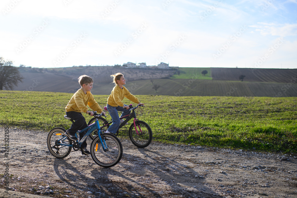 Obraz premium A boy and a girl race their bicycles through the countryside at sunset, laughing and chasing each other. The golden light and peaceful surroundings create a joyful, carefree moment of childhood advent