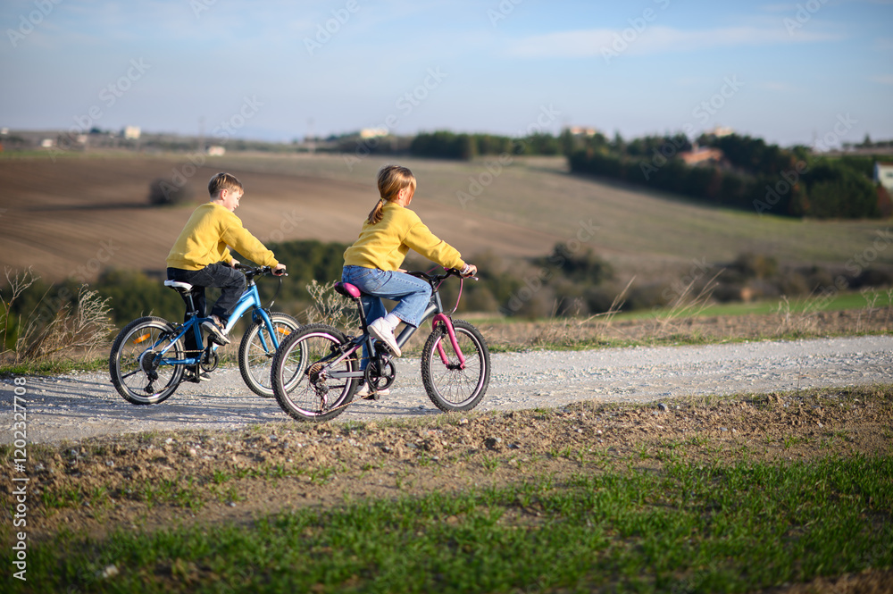 Obraz premium A boy and a girl race their bicycles through the countryside at sunset, laughing and chasing each other. The golden light and peaceful surroundings create a joyful, carefree moment of childhood advent