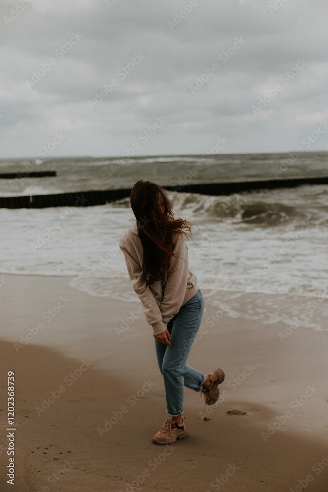 A young girl in a brown sweater and jeans dances on the sandy shore of the sea in windy weather.