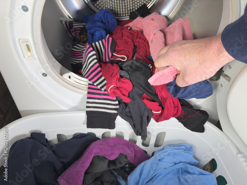 Closeup of a man’s hand pulling a variety of washed and dried clothes from a front loading tumble dryer drum, straight into a laundry basket on the floor. photo