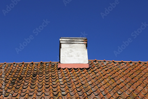 Chimney isolated on blue sky. Old orange ceramic roof tiles. Tiled roof texture. Chimney concrete vent structure at the rooftop. Dirty grunge weathered roofing. Vintage architecture empty copy space. photo