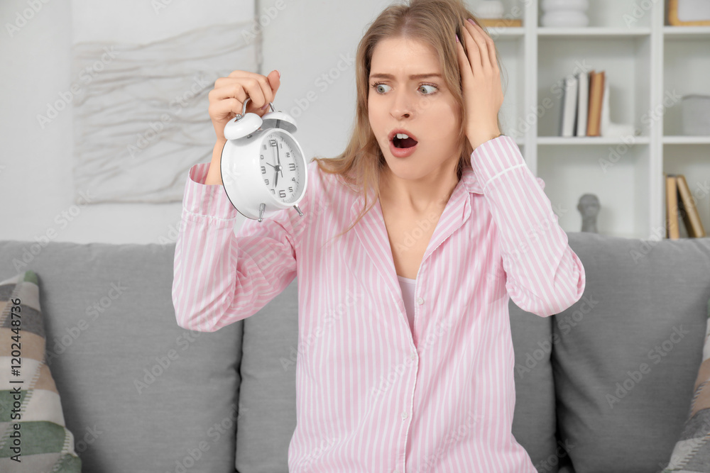 Shocked young woman in pajamas with alarm clock sitting on sofa at home Shocked young woman in pajamas with alarm clock sitting on sofa at home