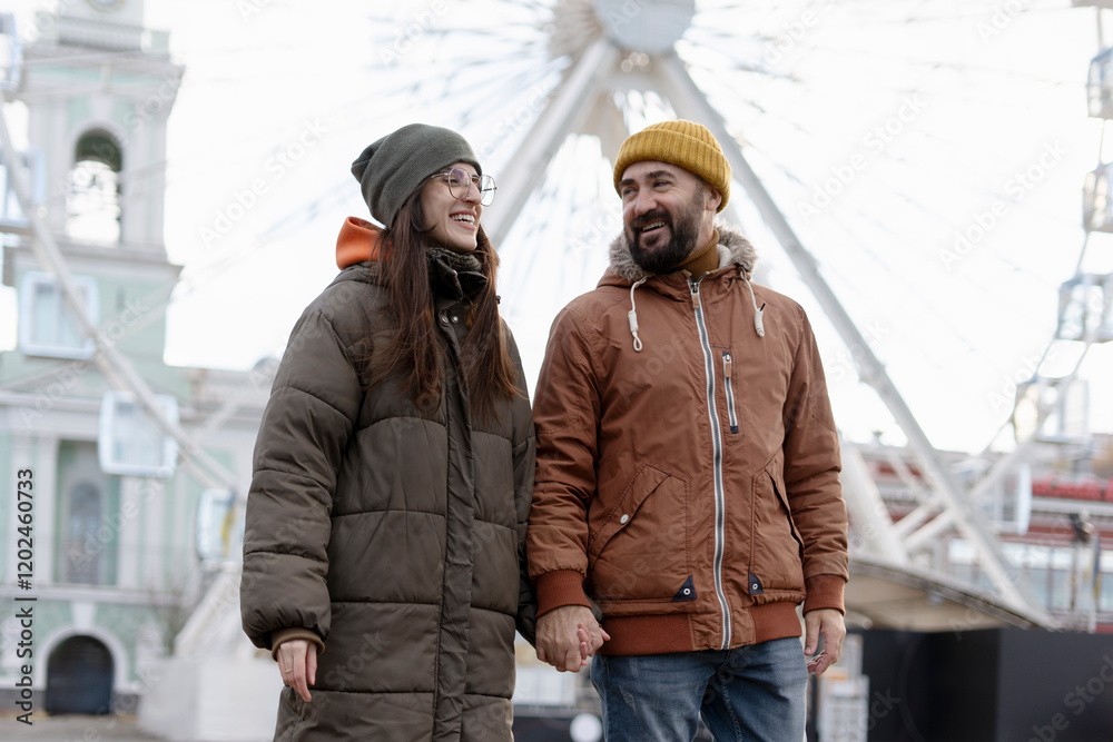 Fototapeta premium Happy couple holding hands and walking near ferris wheel in amusement park