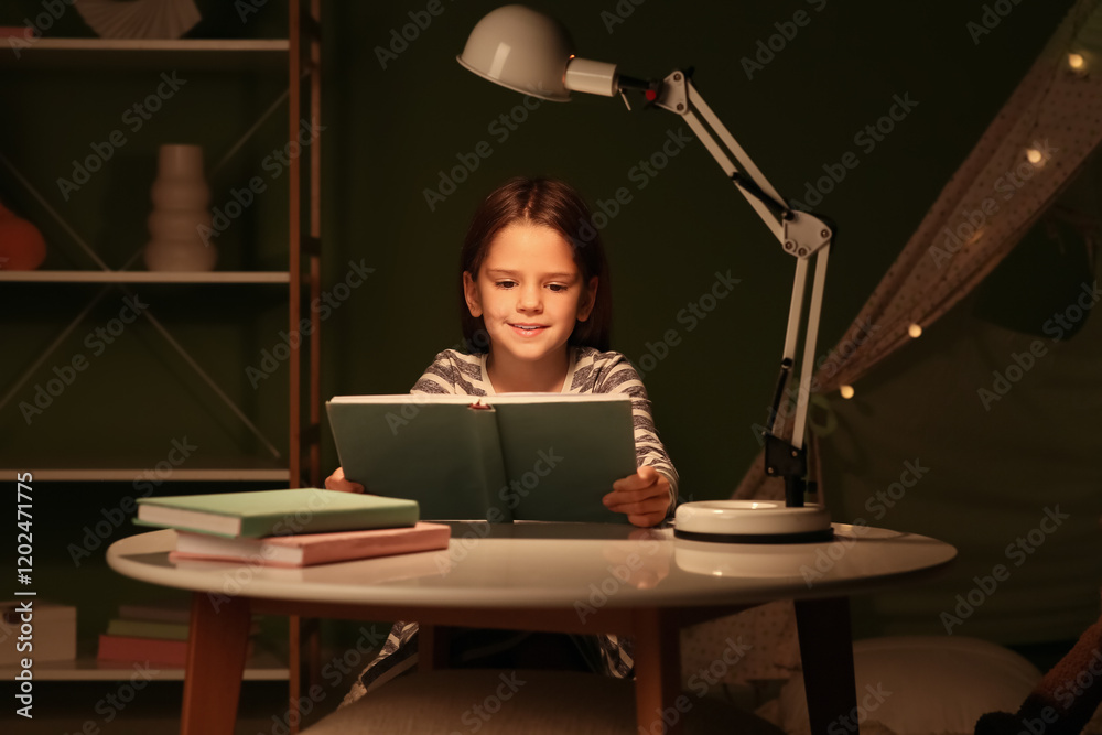 Cute little girl reading book at table with glowing lamp at home Cute little girl reading book at table with glowing lamp at home