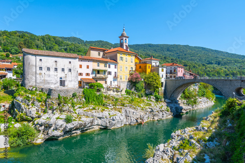 Kanal bridge gracefully stretches over the clear waters of the Soca River, surrounded by charming houses nestled against a backdrop of lush green hills on a sunny day. photo