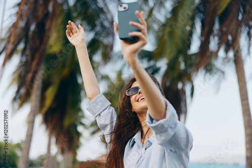 A woman is taking a selfie with her phone while standing in front of a backdrop of numerous lush palm trees, enjoying a tropical vacation in a sunny, beachlike setting photo