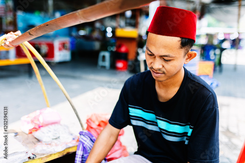 Young street vendor male in songkok hat cooking traditional indonesian food at a market stall, using a wok and charcoal in Jakarta city photo