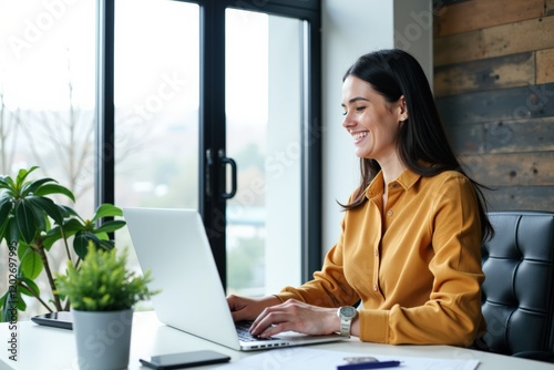 Young Professional Woman Working from Home at a Modern Desk with Laptop, Indoor Plant, and Natural Light Streaming Through a Window photo