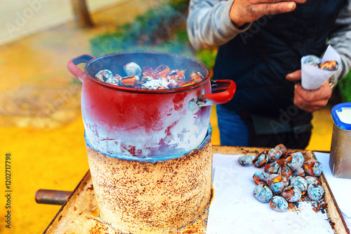 A street vendor roasts chestnuts over a makeshift stove using red pot on a rustic base. Smoky aroma surrounds freshly roasted chestnuts, while cones of paper hold servings for eager customer
