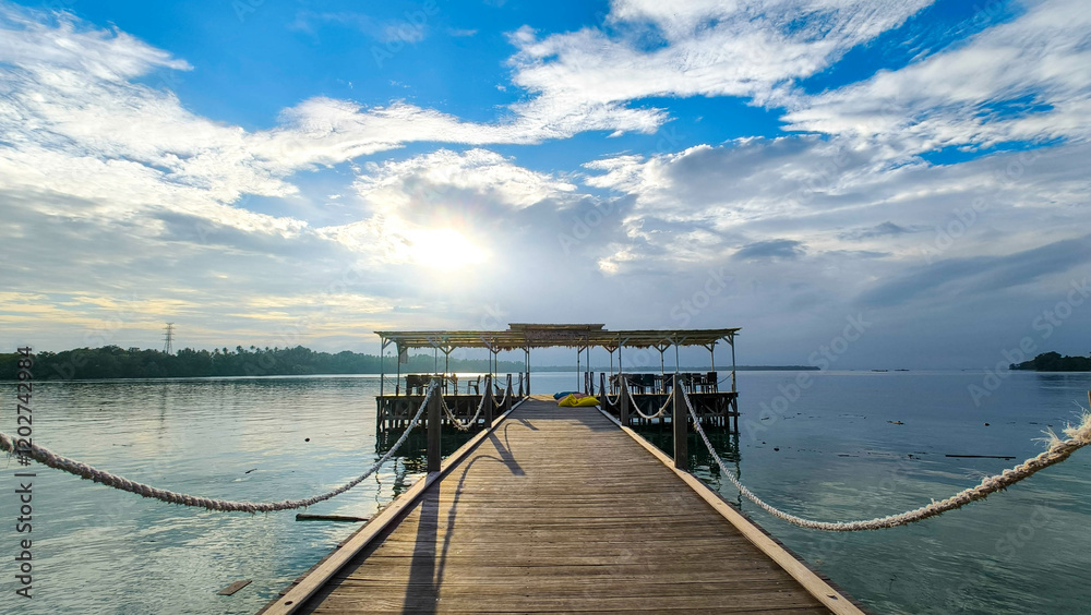 A serene wooden pier extends over calm water, leading to a small structure under a partly cloudy sky with the sun peeking through. The peaceful scene captures the tranquility of nature A serene wooden pier extends over calm water, leading to a small structure under a partly cloudy sky with the sun peeking through. The peaceful scene captures the tranquility of nature