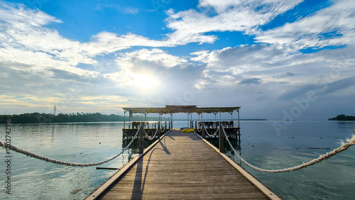 Wallpaper Mural A serene wooden pier extends over calm water, leading to a small structure under a partly cloudy sky with the sun peeking through. The peaceful scene captures the tranquility of nature Torontodigital.ca