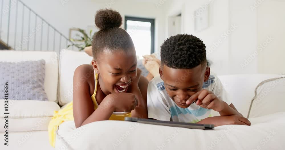 African American siblings lying on couch, watching and laughing at tablet screen African American siblings lying on couch, watching and laughing at tablet screen